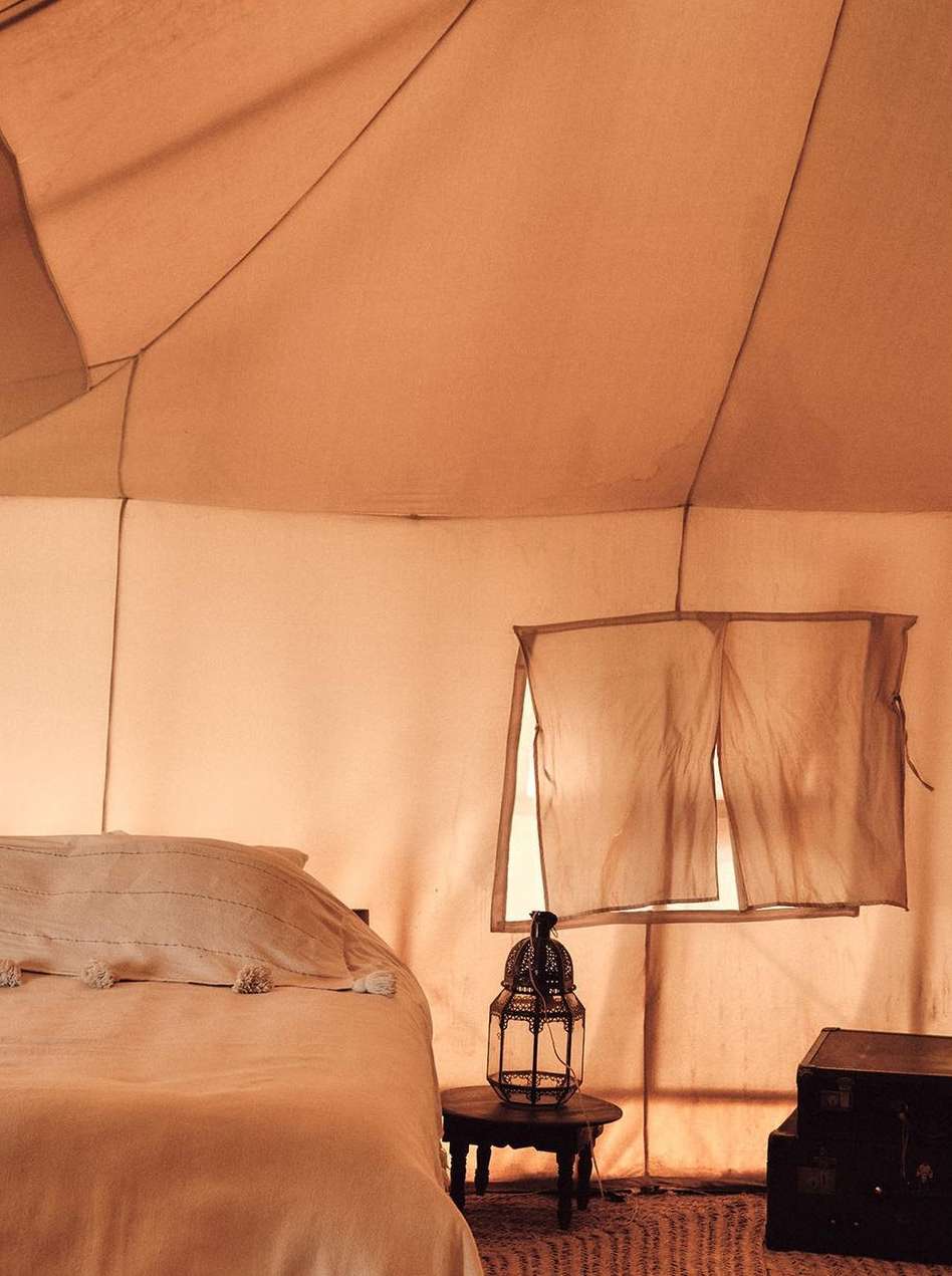 Interior view of a safari-style tent with a bed, bedside table with a lantern, wooden chest, and a rug on the floor.