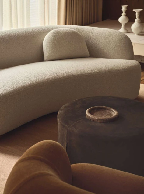Interior design featuring a cream-colored curved sofa, a dark brown round ottoman, and a caramel-colored chair in a neutral color palette.
