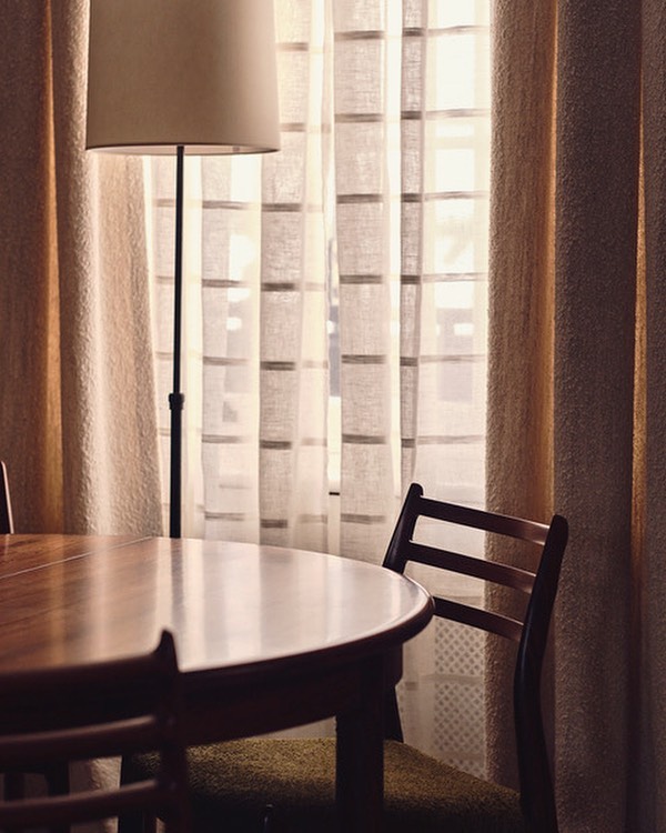 Interior view of a round wooden table and chair set next to a standing lamp with a tan shade and curtains in the background.