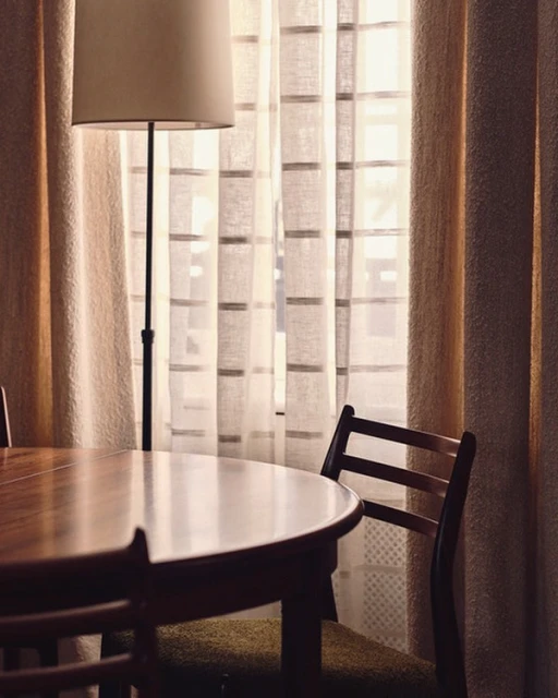 Interior view of a round wooden table and chair set next to a standing lamp with a tan shade and curtains in the background.