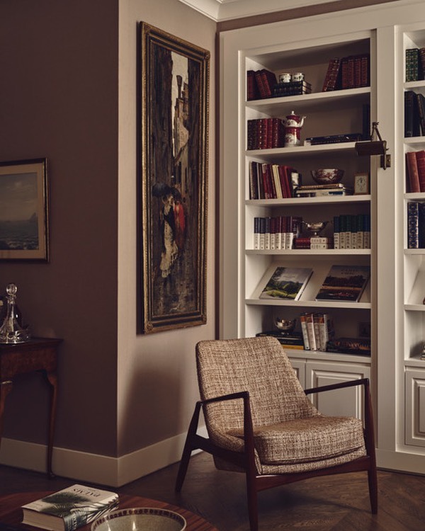 Interior view of a living room with a patterned armchair, a tall bookshelf, and an ornate painting, complemented by warm, soft lighting.