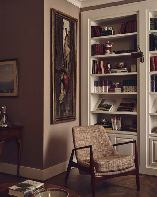 Interior view of a living room with a patterned armchair, a tall bookshelf, and an ornate painting, complemented by warm, soft lighting.