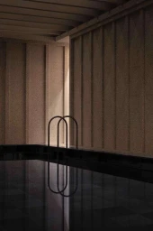 Indoor swimming pool with dark water and metal ladder rails against paneled concrete walls photographed by Ben Anders