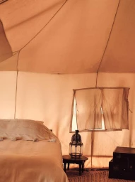 Interior of a canvas tent with a neatly made bed, a small round table holding a lantern, and stacked dark trunks.
