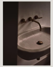 Minimalist concrete bathroom sink with brass wall-mounted faucet and handles in low-key lighting by Ben Anders