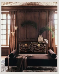 Dark velvet daybed with draped throw against wood paneled wall with floor lamps and vase with palm fronds.