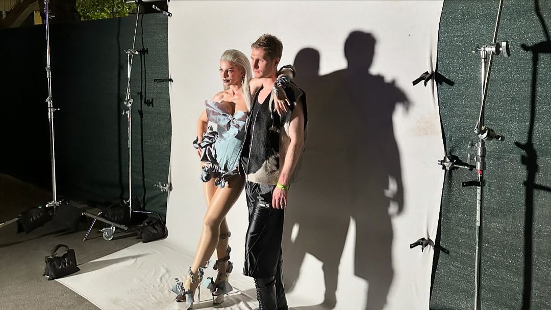 Fashion photo shoot features an adult male and female posing in avant-garde outfits against a white backdrop in a photo studio.