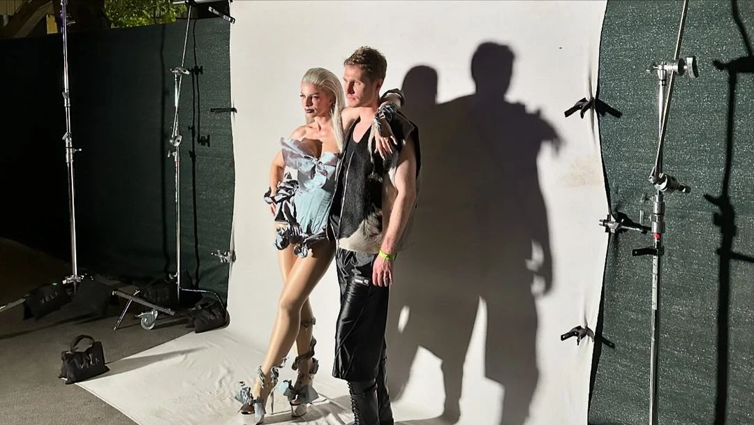 Fashion photo shoot features an adult male and female posing in avant-garde outfits against a white backdrop in a photo studio.