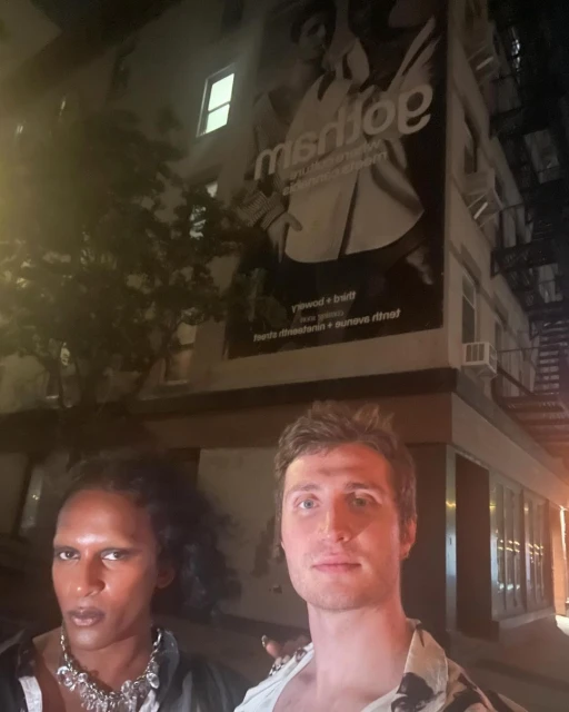 Two adult humans pose at night near a building with an advertisement. One has dark skin, a necklace, and curly hair.