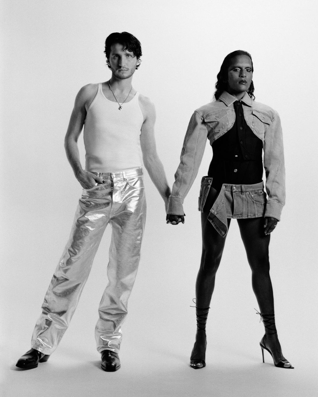 Two people, one male and one feminine-presenting, stand holding hands in a studio wearing futuristic and stylish black and silver outfits.