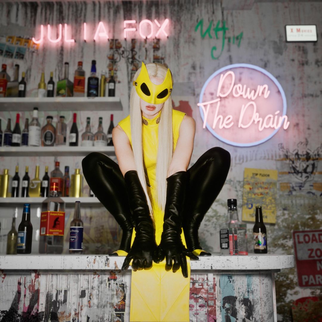 Julia Fox poses in a yellow costume with a mask, sitting on a bar with liquor bottles, against a graffitied wall and neon signs.