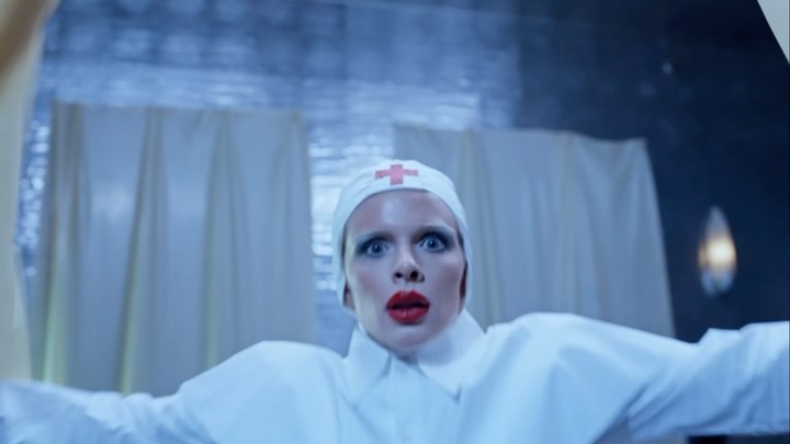 Woman in nurse costume with red lipstick looks at camera indoors with white curtains and a blue-grey background, low angle, medium shot.
