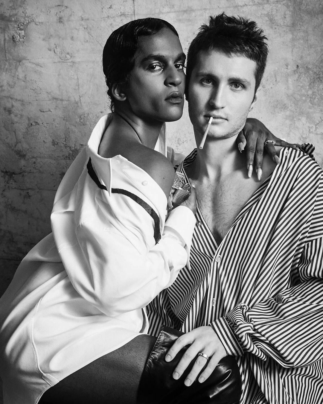 Black and white studio shot of two people with open shirts, one holding a cigarette, against a textured backdrop with soft lighting.