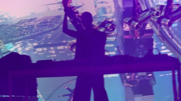 Silhouette of a DJ raising a hand in front of a mixing console with a large screen displaying abstract city visuals in purple light.