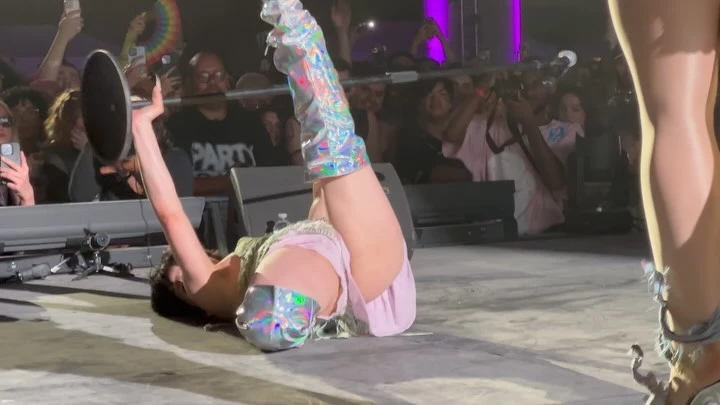 Female performer lies on stage with holographic boots, holding cymbal in front of a crowd, captured with stage lighting.