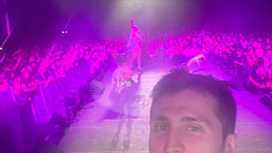 Crowd views a concert with stage performers and colorful lighting while a person takes a selfie from the crowd at a music venue.