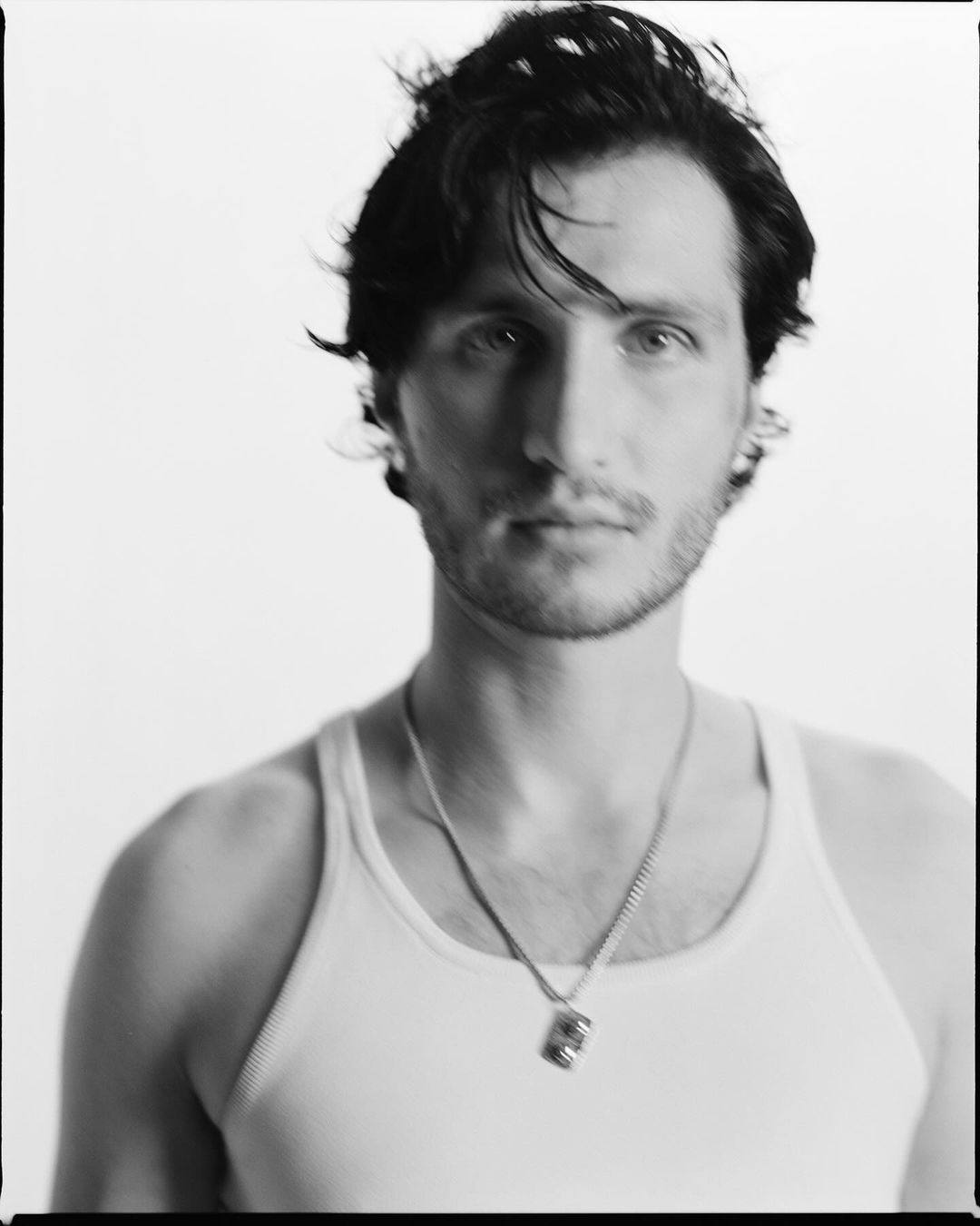 Monochrome portrait of an adult man in a tank top and necklace, shot in a studio setting with a soft, diffused lighting.