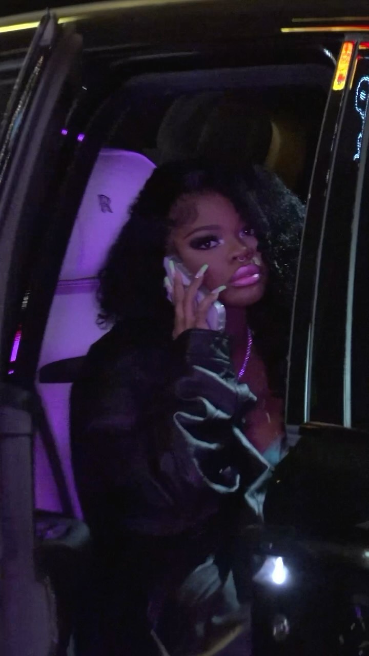 Woman with dark hair and pink lipstick talks on a cell phone in a car with purple lighting and light colored interior.