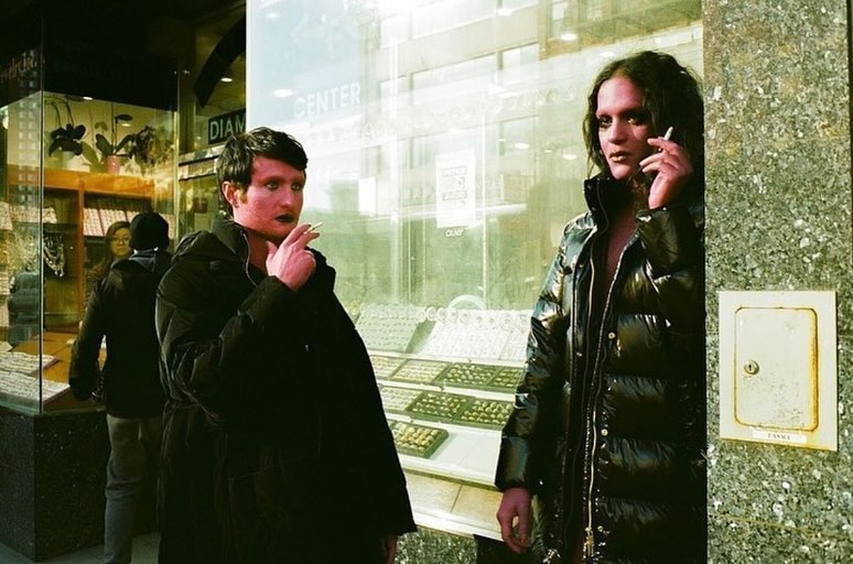 Two people stand outside a jewelry store, one smoking, the other on a phone. Both are wearing dark clothing and are positioned near a shop window.