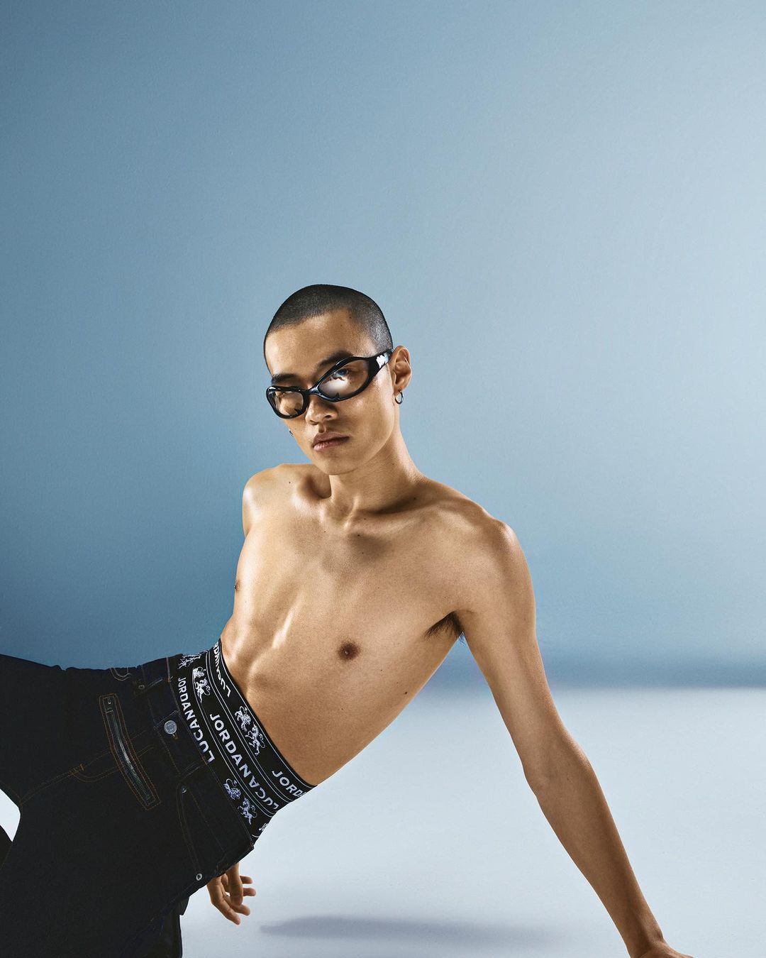 Shirtless man wearing dark sunglasses and branded black jeans against a blue studio background