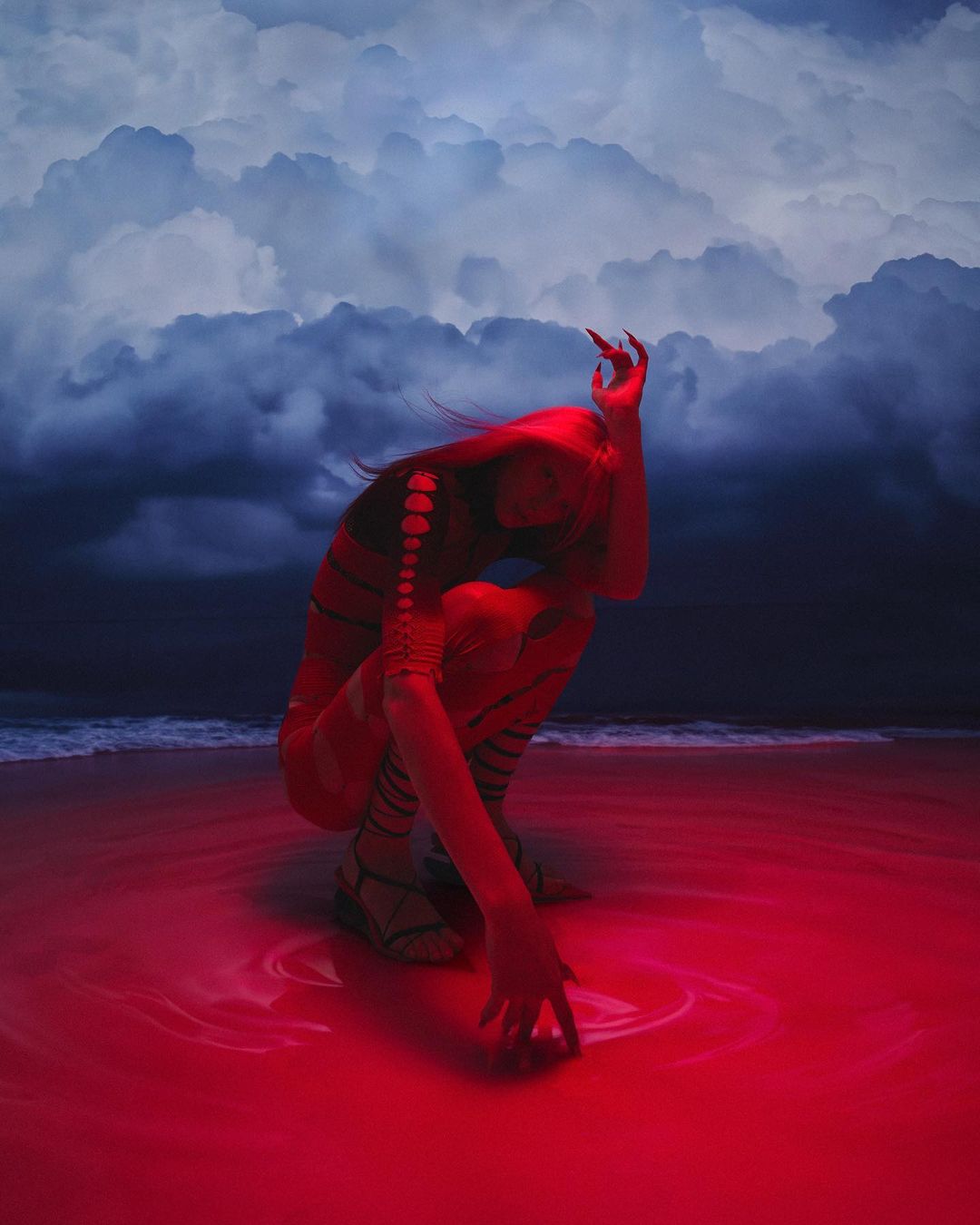 A woman in a red bodysuit crouches on a beach with moody clouds, creating an artistic and surreal image with dramatic lighting and color.