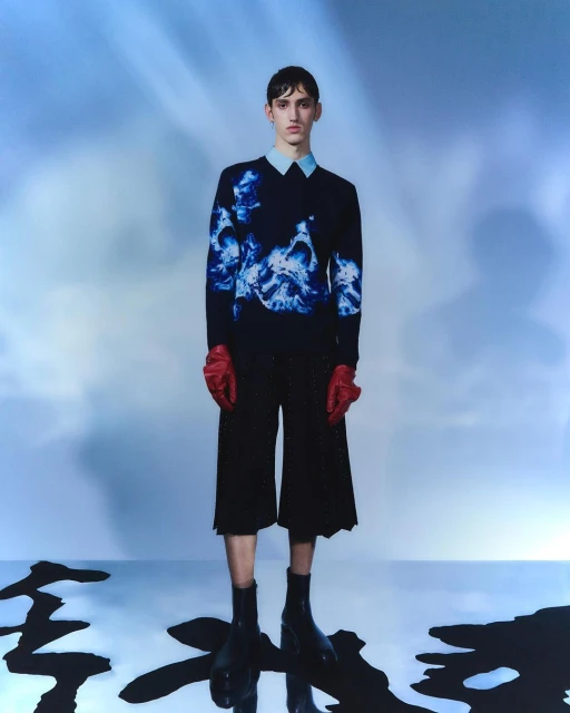 Full-body studio shot of a young adult male fashion model wearing a sweater with a blue print, red gloves, black culottes and black boots.