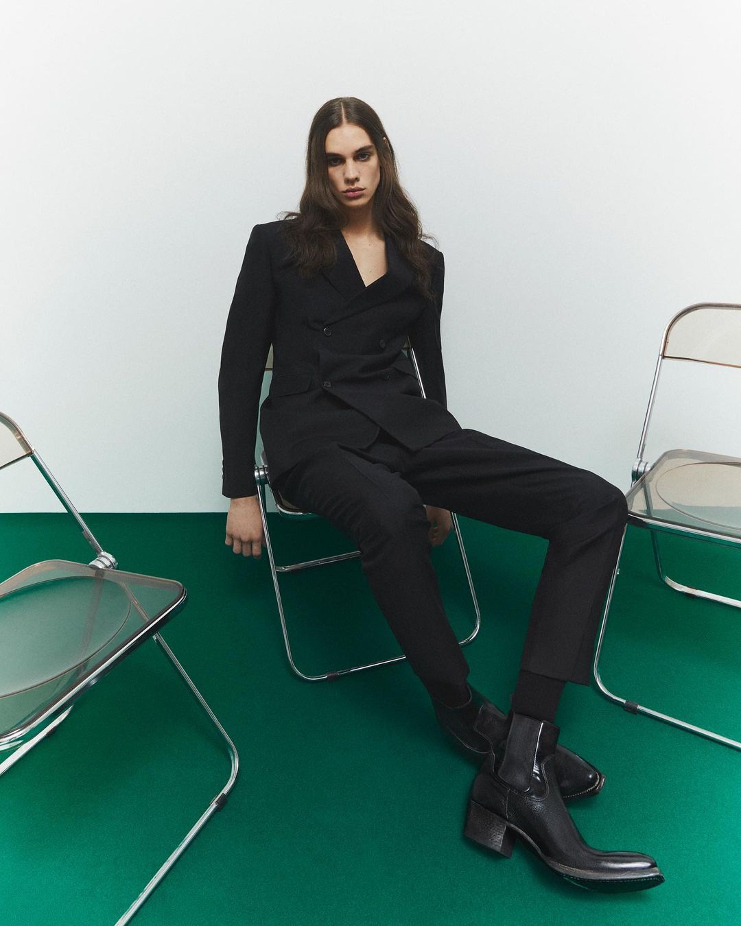 Person in black suit sitting on chair in a studio setting. Dark hair and black boots complete the look. Fashion photography.