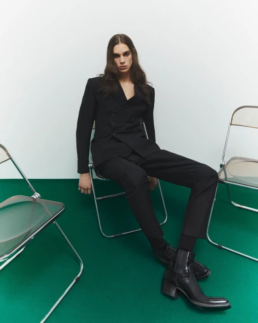 Person in black suit sitting on chair in a studio setting. Dark hair and black boots complete the look. Fashion photography.
