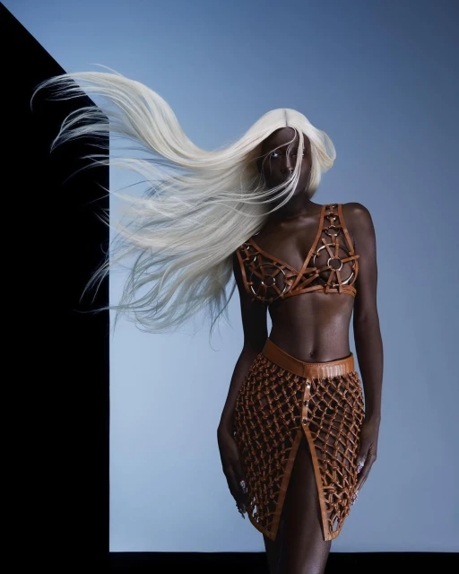 Woman with platinum blonde hair wearing a brown leather bra top and skirt against a blue and black background in a studio.