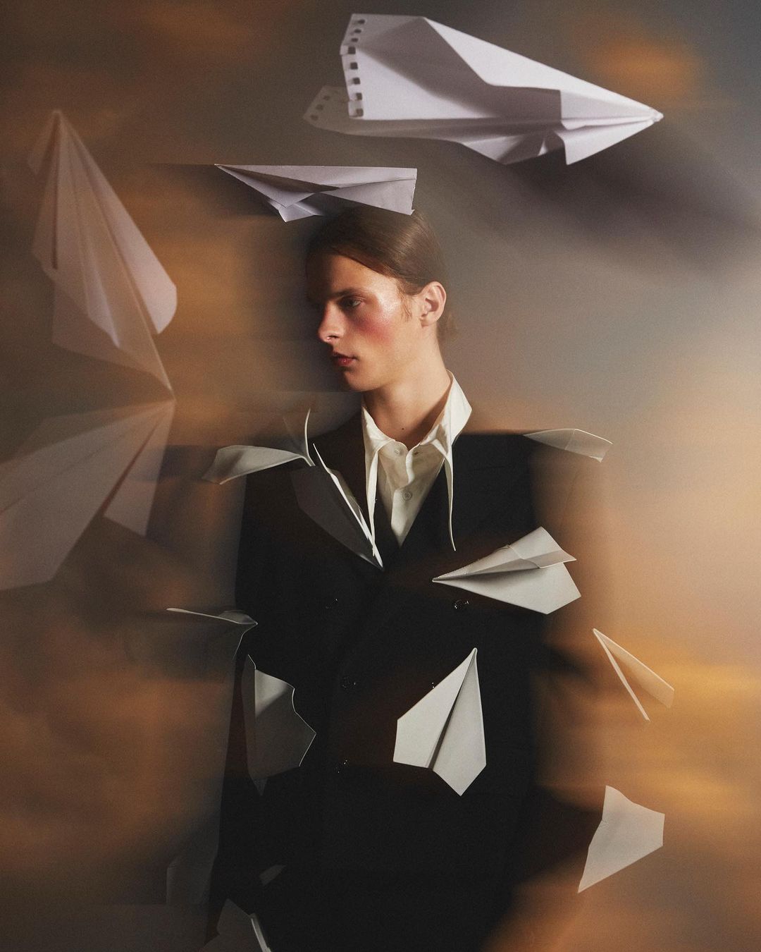 Artistic studio shot of a young adult in a suit jacket surrounded by paper airplanes with a soft orange and grey background.