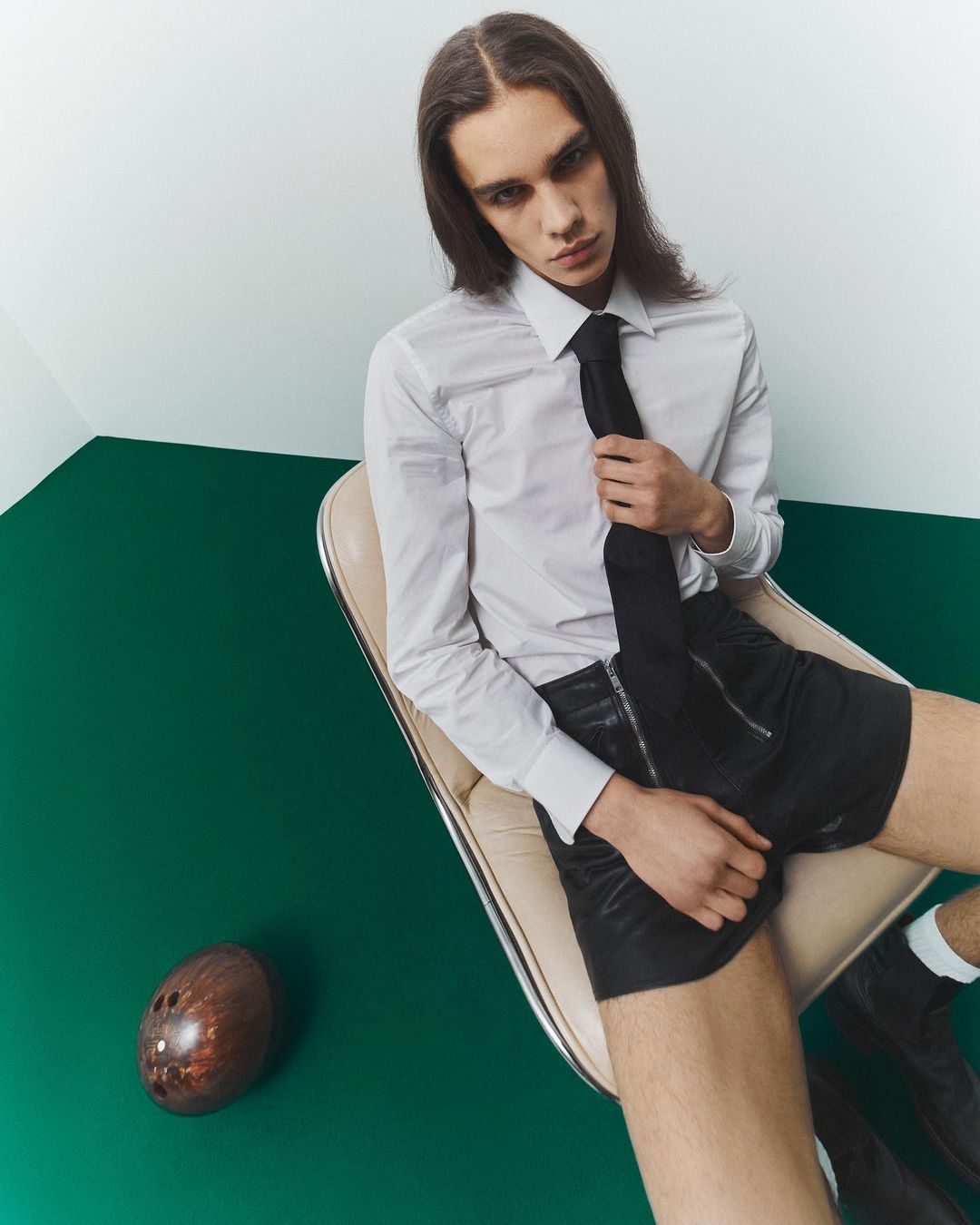 A slender man with long brown hair poses in a white shirt, black tie, and shorts while sitting in a chair with a football on a green floor.