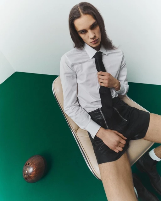 A slender man with long brown hair poses in a white shirt, black tie, and shorts while sitting in a chair with a football on a green floor.