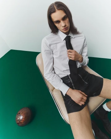 A slender man with long brown hair poses in a white shirt, black tie, and shorts while sitting in a chair with a football on a green floor.