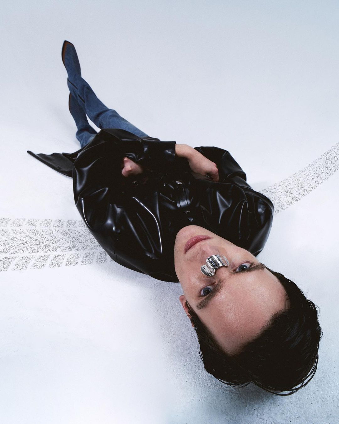 Adult male in a black trench coat and blue jeans lies on a white surface with a silver face accessory looking into the camera.