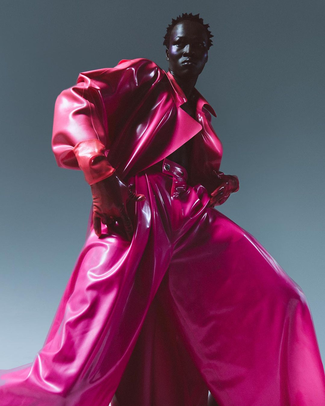 Fashion photo of a Black adult female model in a vibrant magenta and orange shiny trench coat and wide-leg pants, posing in a photo studio.