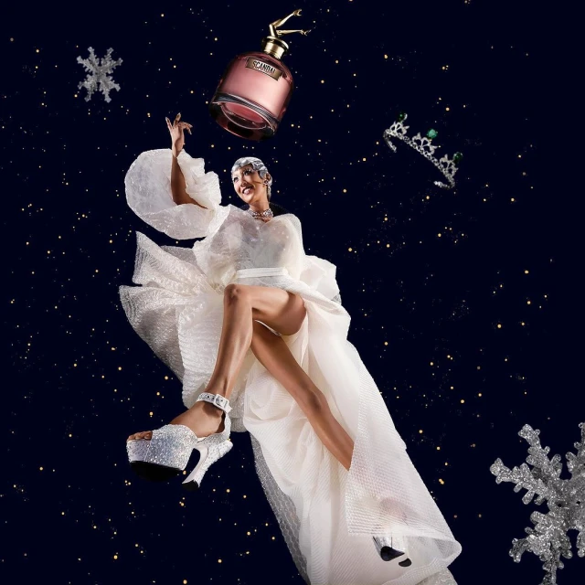 A woman in a white dress with a perfume bottle and tiara floating above her against a blue star field background.