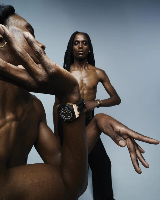 Two dark-skinned adult males strike dynamic poses in a photo studio. One wears a watch, with dramatic lighting against a gradient grey backdrop.