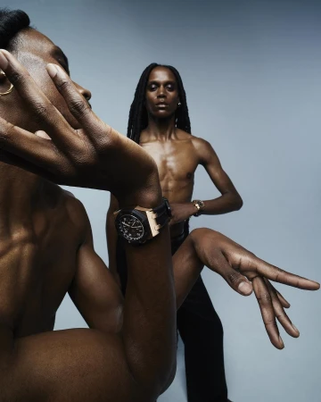 Two dark-skinned adult males strike dynamic poses in a photo studio. One wears a watch, with dramatic lighting against a gradient grey backdrop.