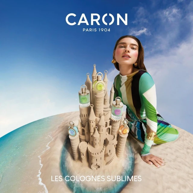 Caron Paris perfume bottles are arranged in a sandcastle formation on a beach, with a woman in a colorful dress in the background.