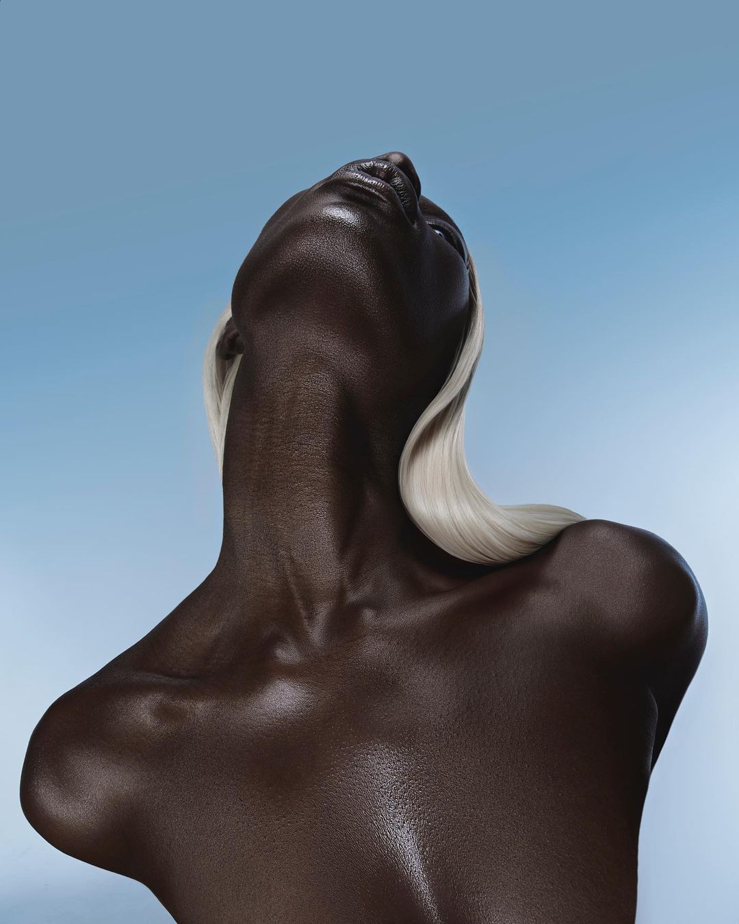 Striking portrait of a dark-skinned woman with platinum blonde hair, head tilted back, set against a clear, light blue sky backdrop.