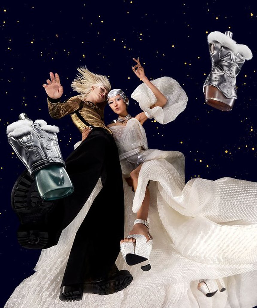 Two models wearing avant-garde fashion pose against a dark blue starry backdrop, showcasing futuristic styles and accessories. 