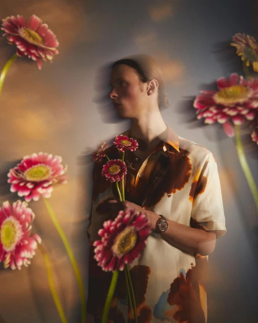 Man in patterned shirt holds pink Gerbera daisies surrounded by more daisies in a softly lit, artistic portrait.