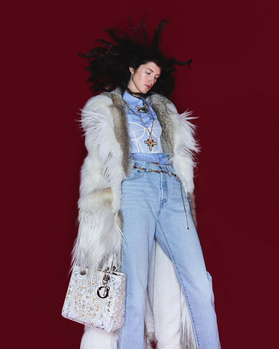 Fashionable woman in a fur coat, light blue jeans, and carrying a Dior bag against a red background, showcasing high-fashion style.