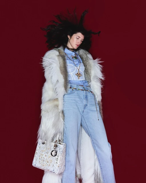 Fashionable woman in a fur coat, light blue jeans, and carrying a Dior bag against a red background, showcasing high-fashion style.