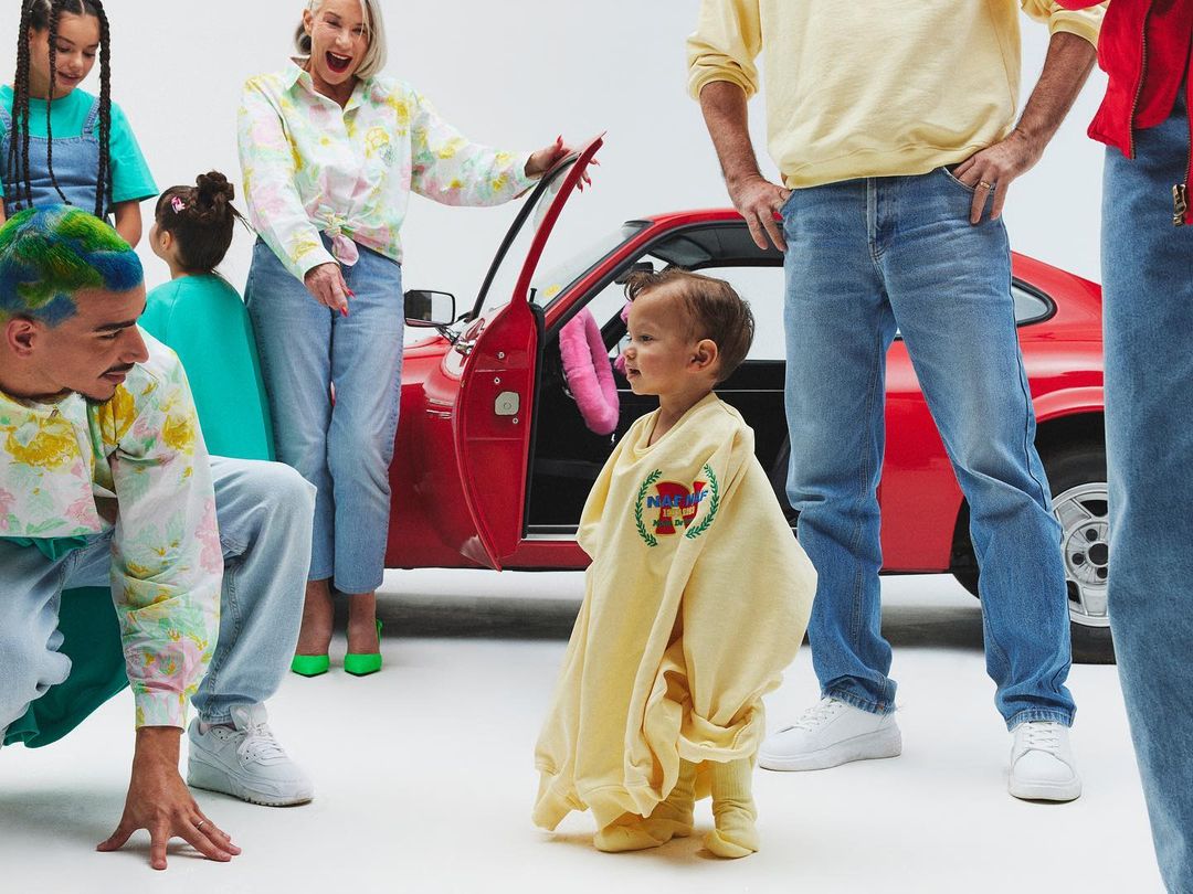 A group of people of various ages stand near a vintage red car. A baby in an oversized yellow sweatshirt stands in the foreground.