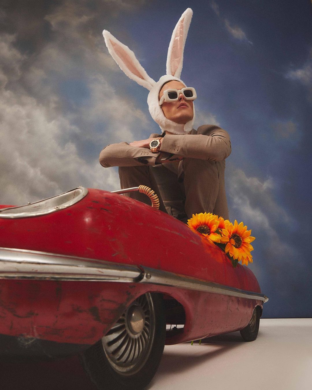 Person in bunny hat and sunglasses seated in a red toy car with sunflowers, against a cloudy sky backdrop in a fashion editorial.