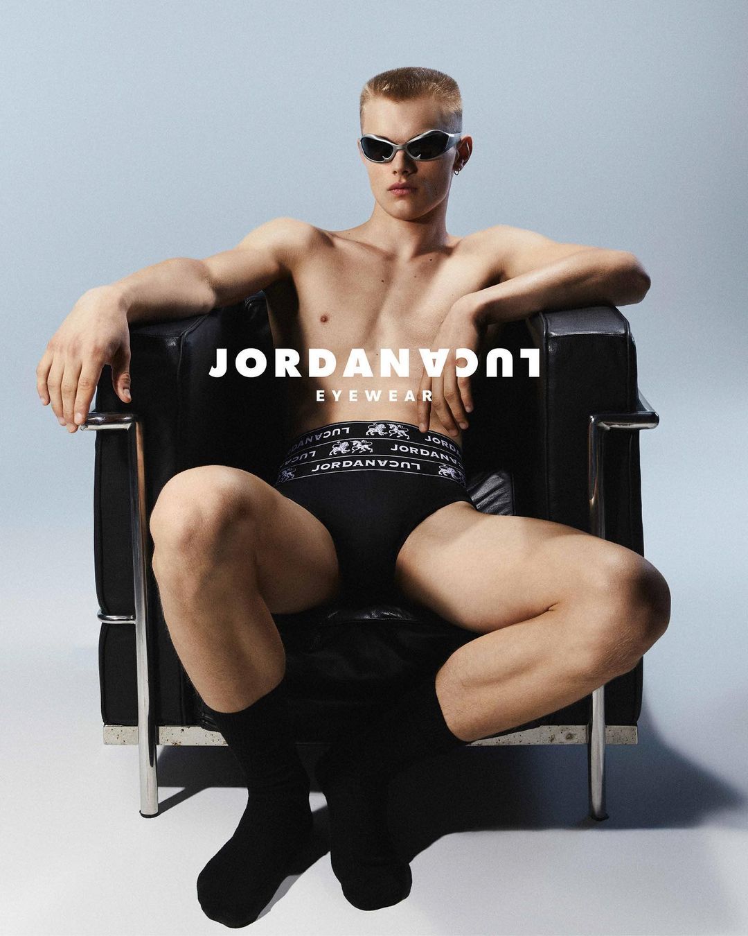 A shirtless adult male wearing sunglasses sits in a black chair against a light blue background in a studio shot for an eyewear advertisement.