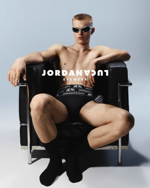 A shirtless adult male wearing sunglasses sits in a black chair against a light blue background in a studio shot for an eyewear advertisement.