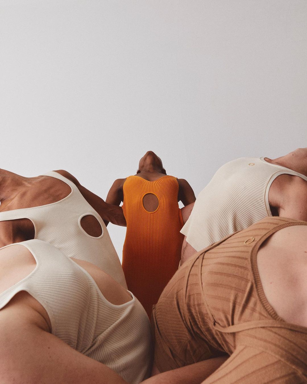 Five women in minimalist dresses are arranged in a circular formation against a stark white backdrop in a studio shot.