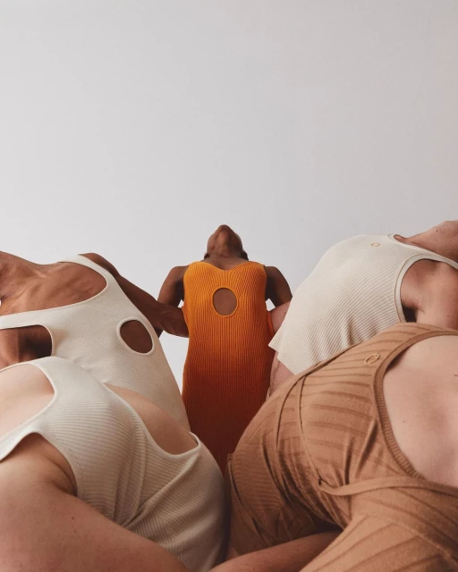 Five women in minimalist dresses are arranged in a circular formation against a stark white backdrop in a studio shot.